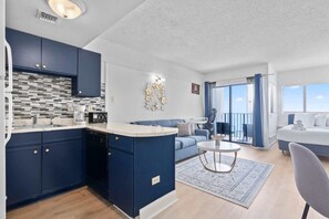 Une cuisine moderne avec des meubles de couleur bleu foncé, un plan de travail blanc et un pare-feu. On y trouve un coin repas avec un canapé bleu et une table ronde. Un balcon avec des rideaux bleus est visible.