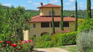 A two-story building with a tiled roof, surrounded by lush greenery and tall cypress trees.