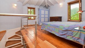 A bedroom with a bed, a wooden staircase, a wardrobe, and a window with a view of greenery.