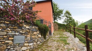 A stone wall with a metal gate, a wooden fence, and a path leading to a building with pink flowers.
