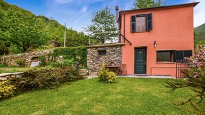 A two-story house with a stone wall and a red exterior. There is a small garden with a lawn and a few plants.