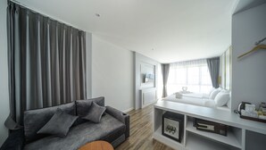 Ein modernes Hotelzimmer mit grauem Sofa, einem kleinen Holztisch, einem weißen Schminktisch mit Spiegel und einem großen Fenster mit Vorhängen.