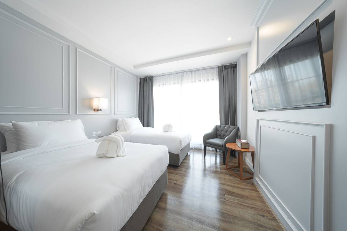 A hotel room with two beds, a grey armchair, a small table, a flat-screen TV mounted on the wall, and a window with curtains.