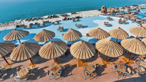 Zona de piscina con sillones de descanso y sombrillas, rodeada por una playa y vistas al agua.