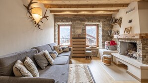 A cozy living room with a stone fireplace, a grey sofa with patterned cushions, and a wooden ceiling. - Casa Ladinia (San Vito di Cadore)