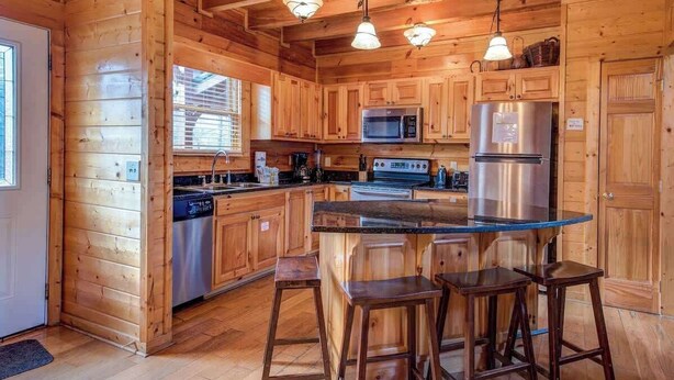 A wooden cabin interior with a kitchen featuring a central island, bar stools, and built-in appliances.