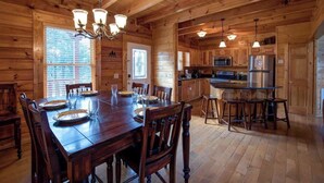 A wooden cabin interior with a dining table set for four, a kitchen area with modern appliances, and a chandelier.