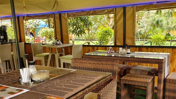 An outdoor dining area with wicker furniture and glass-top tables, surrounded by large windows offering views of greenery.