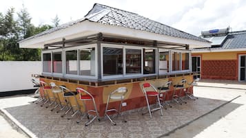 Un pequeño café al aire libre con techo de tejas, zona cubierta con mesas y sillas de metal.