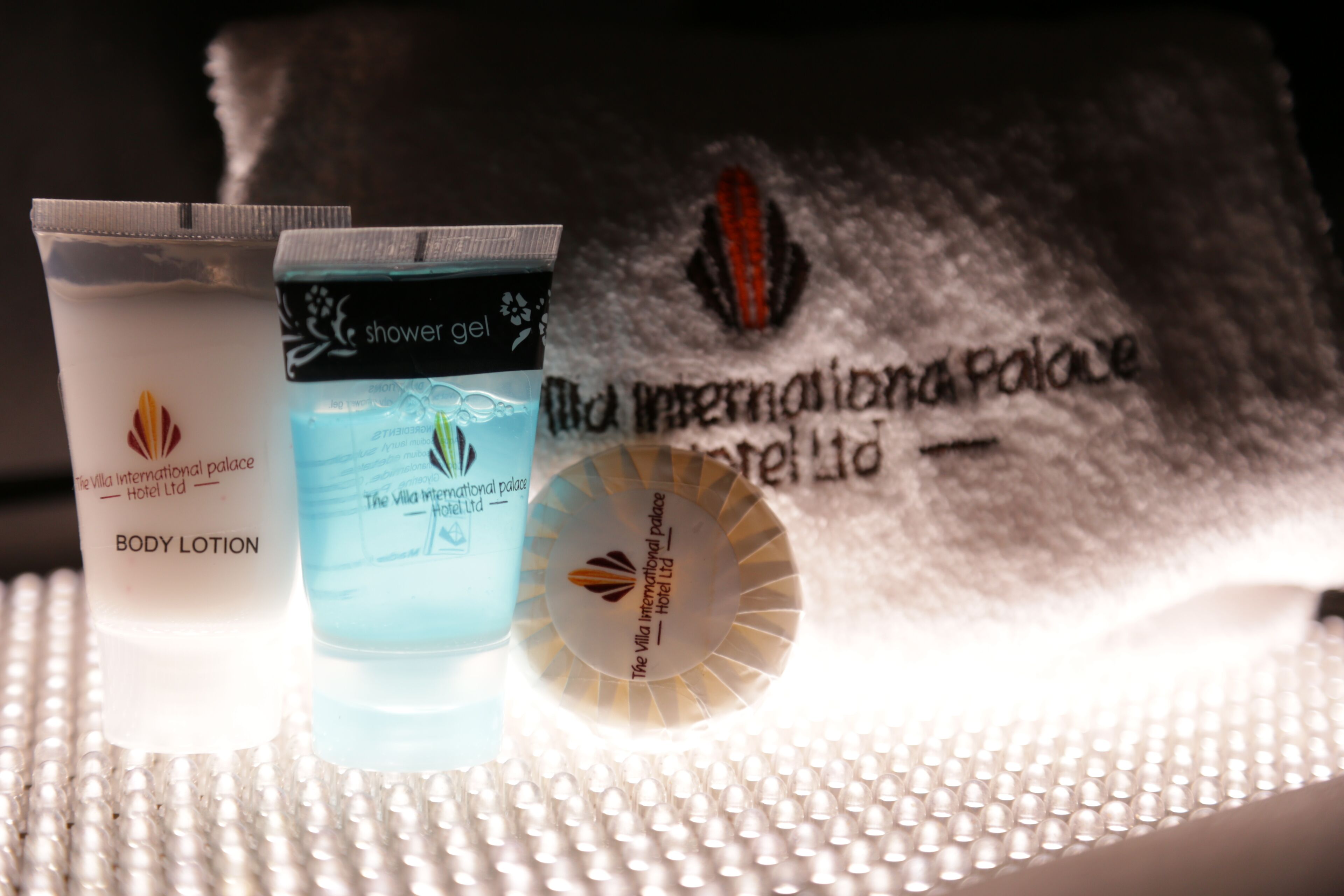 Toiletartikelen, waaronder bodylotion en douchegel, en een handdoek met het logo van Villa International Palace.