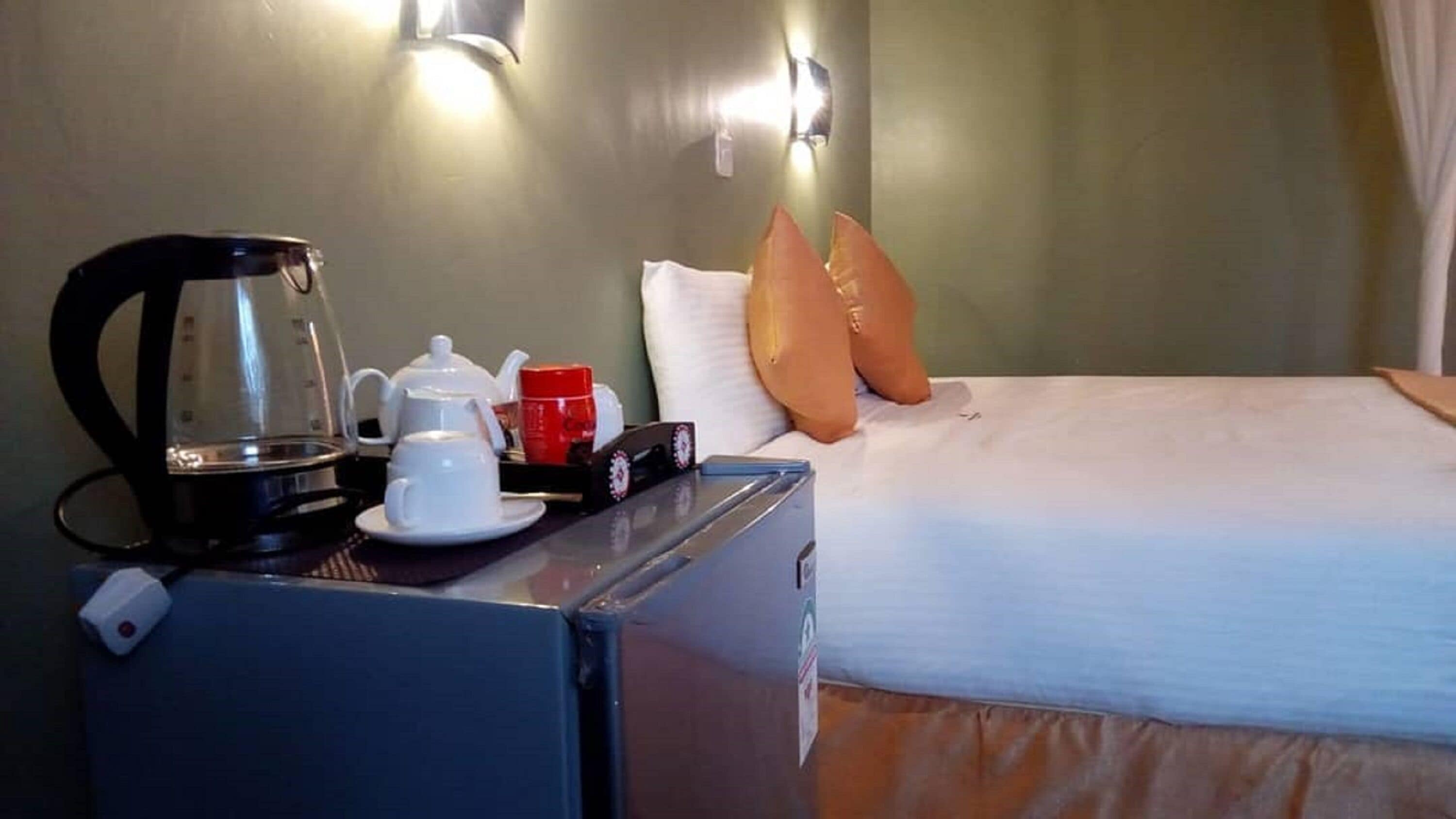 Une chambre d’hôtel avec un lit, une table de chevet sur laquelle se trouvent une bouilloire, une théière, une tasse rouge et un oreiller.