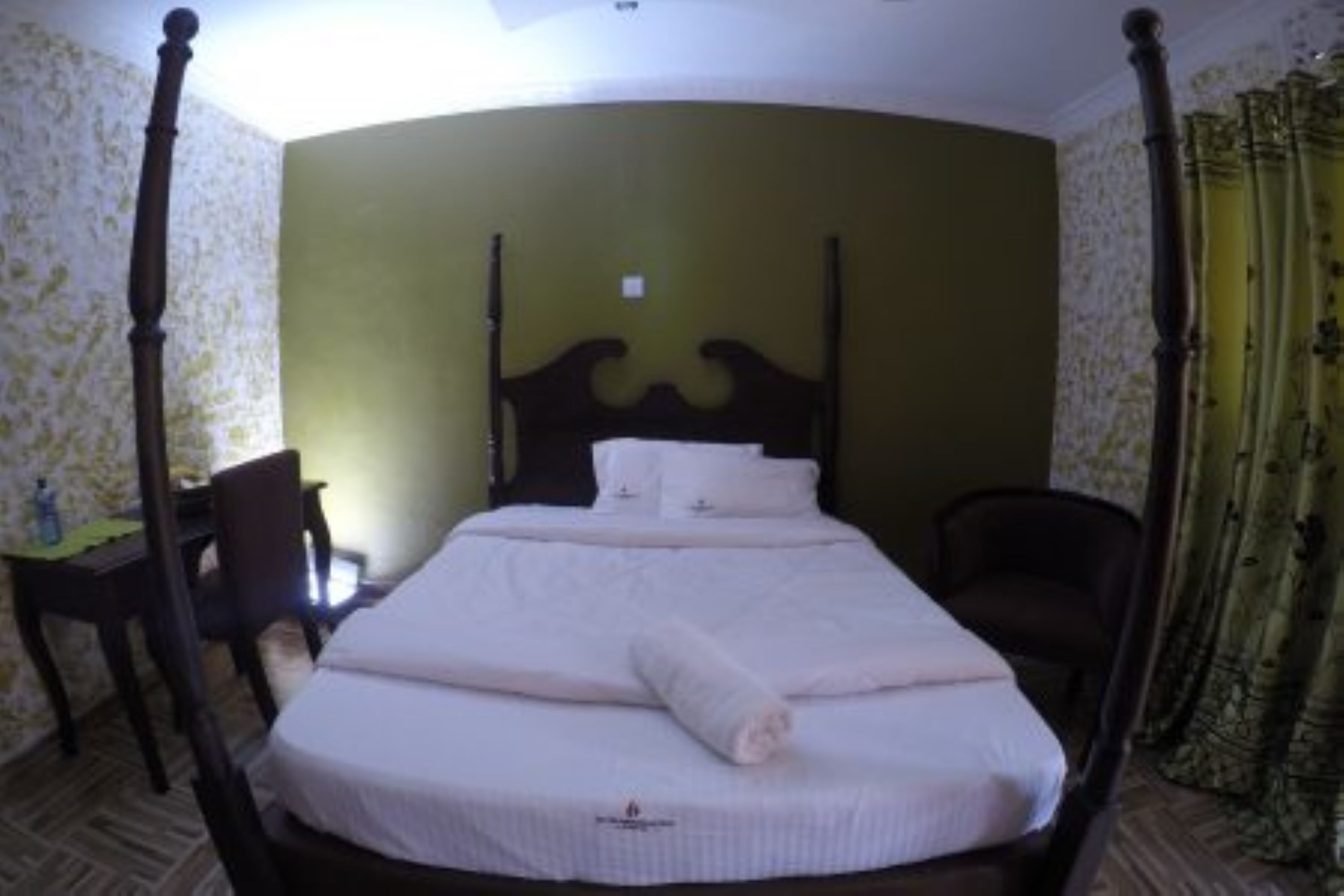 A four-poster bed with white linens and a rolled towel, a wooden desk with a chair, and a green wall with floral wallpaper.