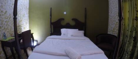 A four-poster bed with white linens and a rolled towel, a wooden desk with a chair, and a green wall with floral wallpaper.