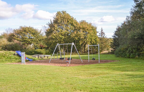 Un parc infantil amb cadira de balanç i tobogan, envoltat d'herba i arbres.