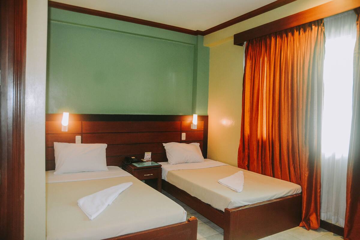 Hotellihuone, jossa on kaksi erillistä sänkyä, puiset päätylevyt, yöpöytä lampun kanssa ja ikkuna, jossa on oranssit verhot.