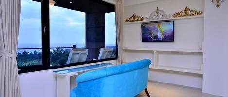 A modern living room with a large window, a blue sofa, a white desk, and a television on the wall.