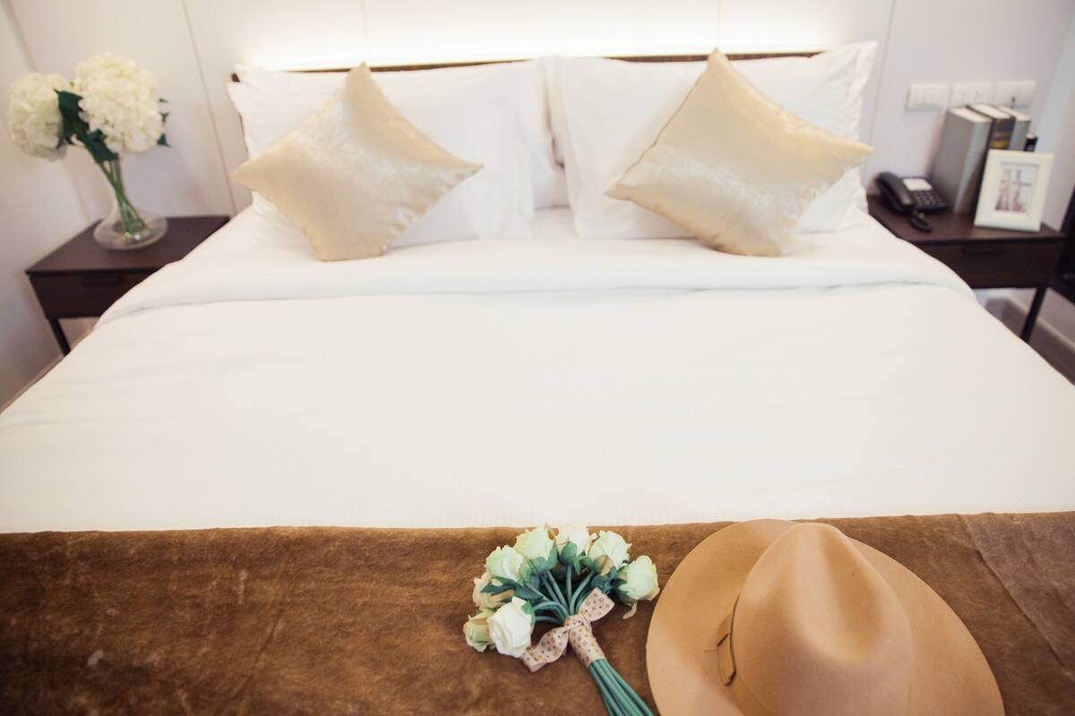 Un lit bien fait, avec du linge de lit blanc et deux oreillers beige, une tête de lit marron et un bouquet de fleurs blanches posé sur la couverture.