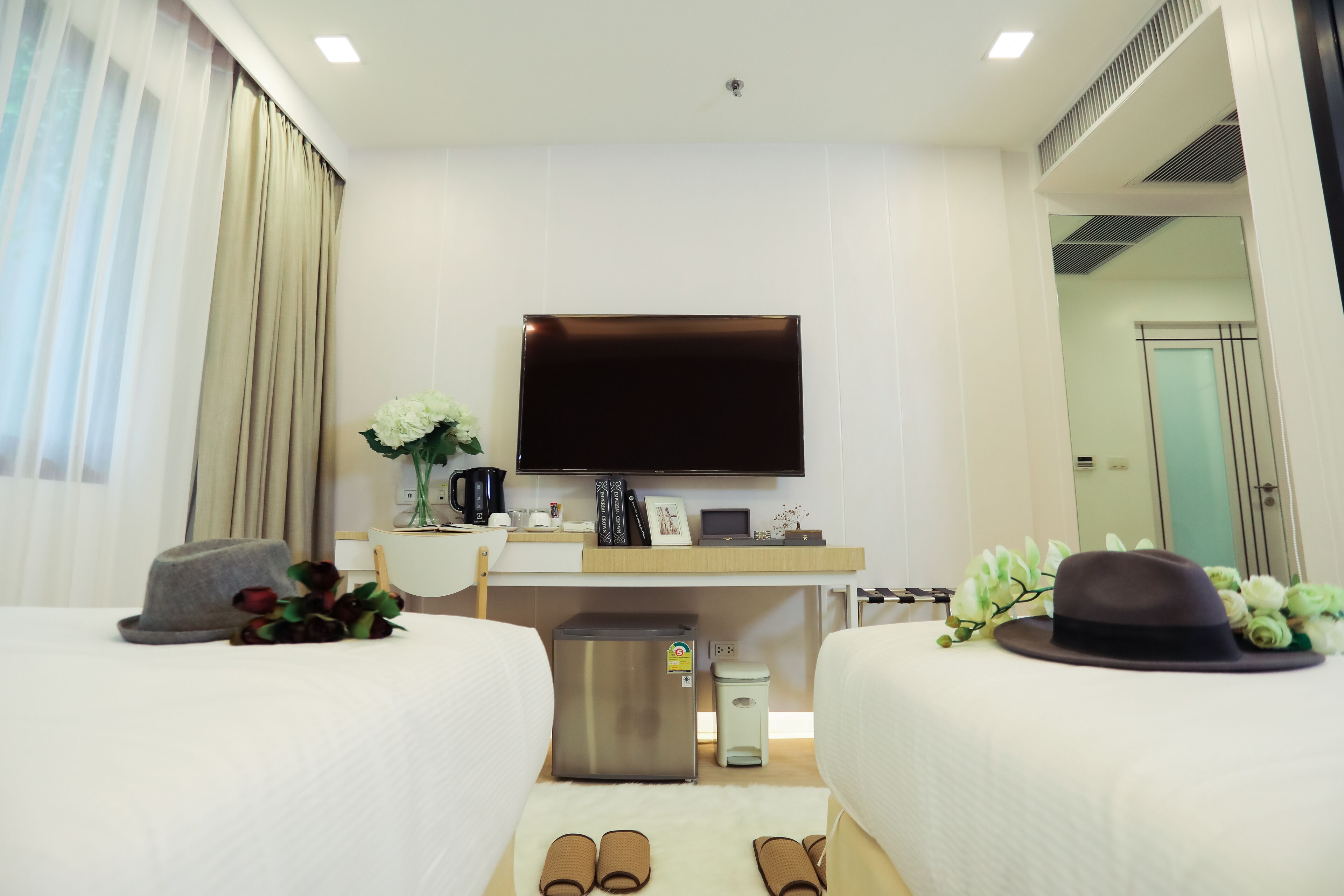 A hotel room with two beds, a TV, a desk with a computer, a small fridge, and a trash bin.