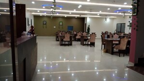 A spacious hotel lobby with multiple dining tables and chairs, a reception desk, and decorative ceiling lights.