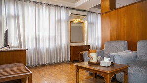 Quarto com piso de madeira, sofá, mesa com cafeteira e lenços de papel, espelho e televisão sobre um suporte.