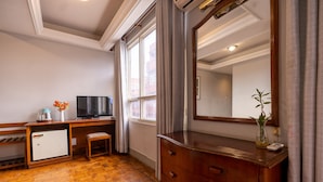Quarto de hotel com piso de madeira, uma escrivaninha com televisão, cômoda e um espelho grande.