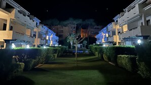 Ein nächtlicher Blick auf ein Hotel mit symmetrischer Architektur, das mehrere Etagen und Balkone aufweist, umgeben von gepflegten Hecken und einer zentralen Palme.