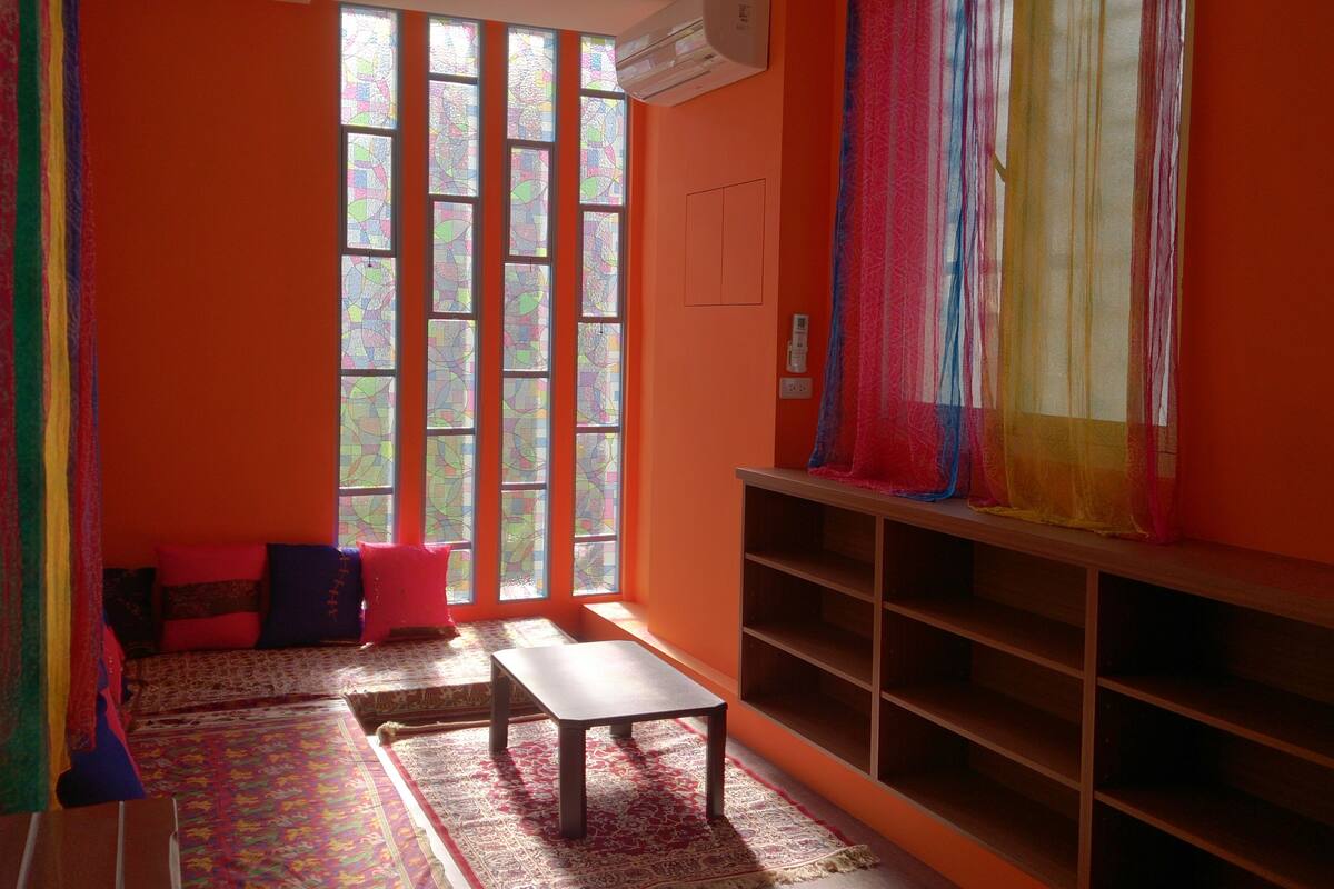 Una habitación con paredes anaranjadas, un ventanal con vidrieras, un banco de madera, una pequeña mesa y una estantería.