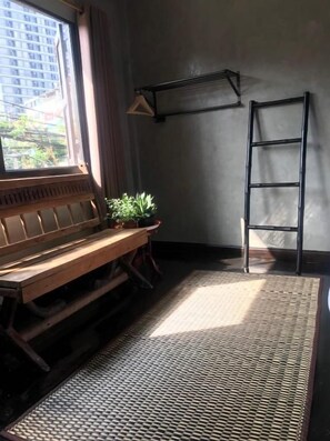A room with a wooden bench, a small potted plant, a ladder, and a window with a view of buildings outside. - Tang House (Bangkok)