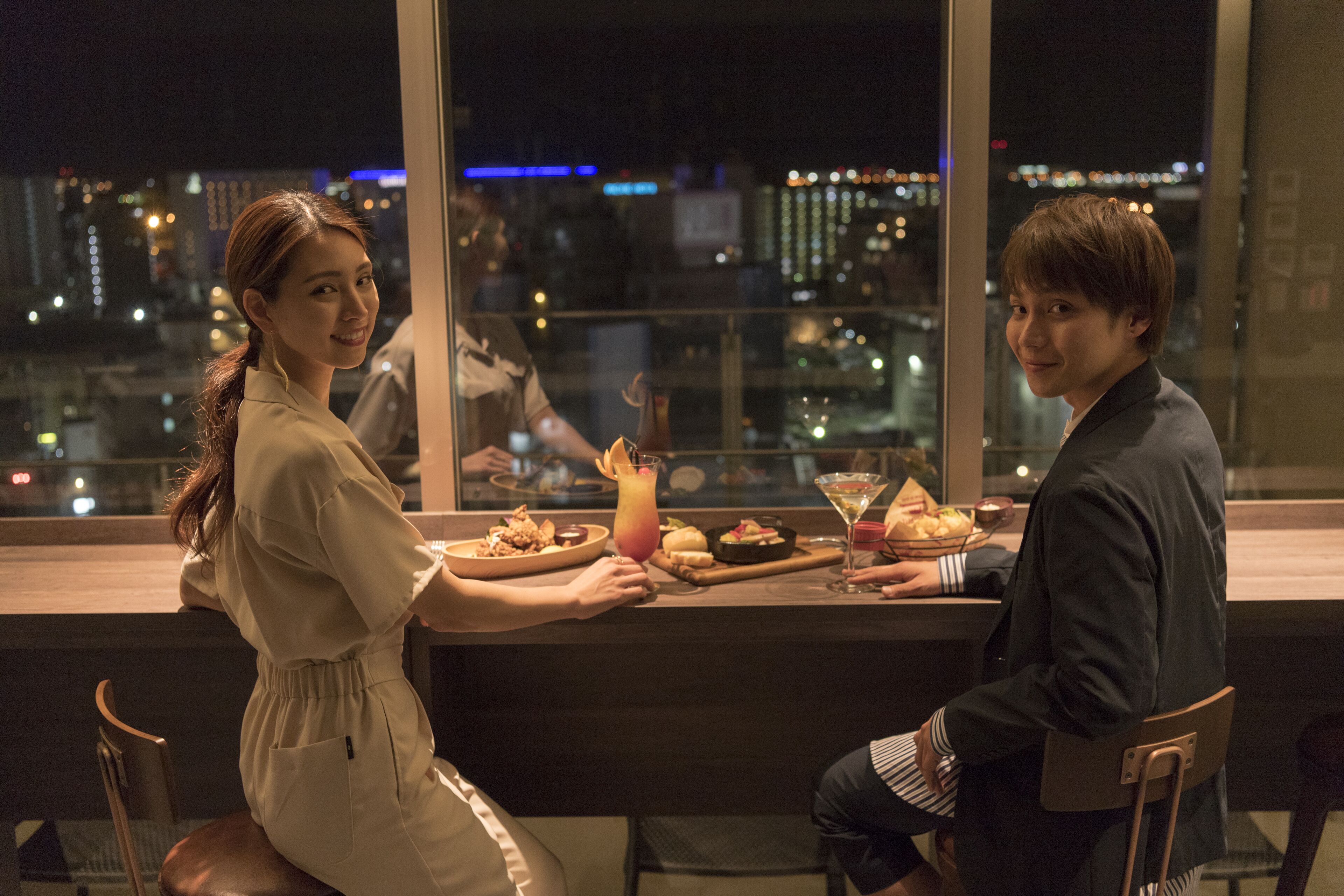 Comedor con vista a la ciudad, una mesa con comida y bebidas, y dos personas sentadas.