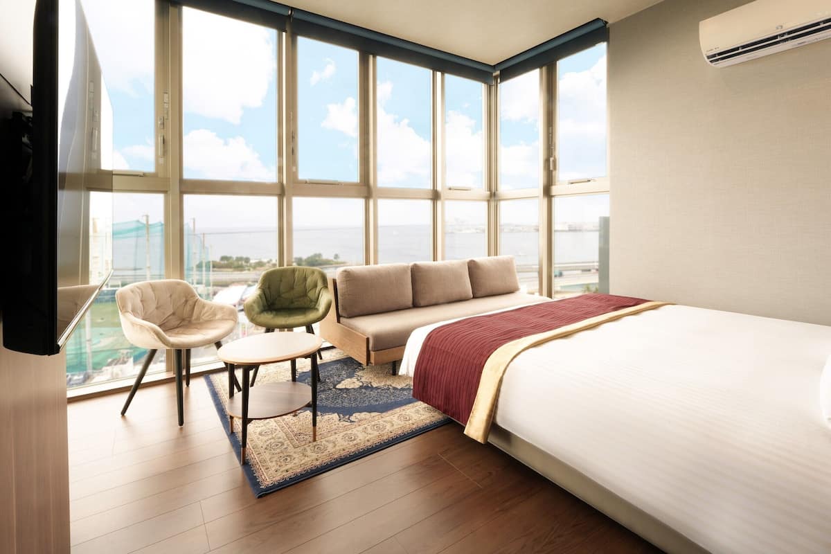 Quarto de hotel com uma cama grande, um sofá, uma cadeira e uma mesa pequena. Há uma janela ampla com vista para a cidade.