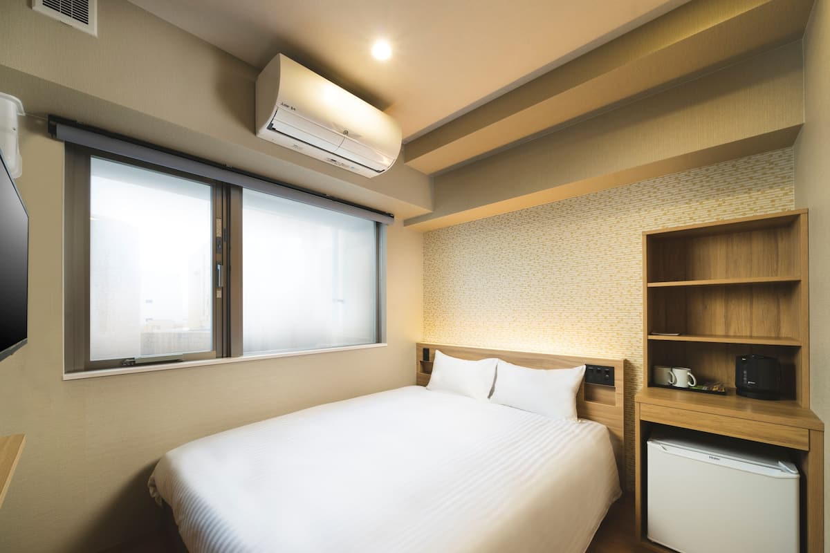 Quarto de hotel com cama, prateleira embutida com cafeteira, mini-geladeira e ar condicionado.