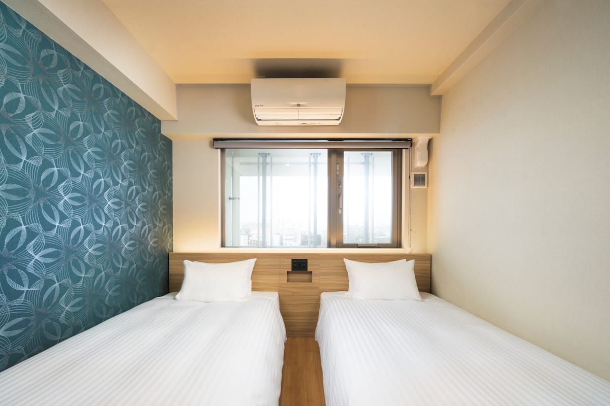 Quarto de hotel com duas camas, uma janela com cortinas, aparelho de ar condicionado e parede com padrão.