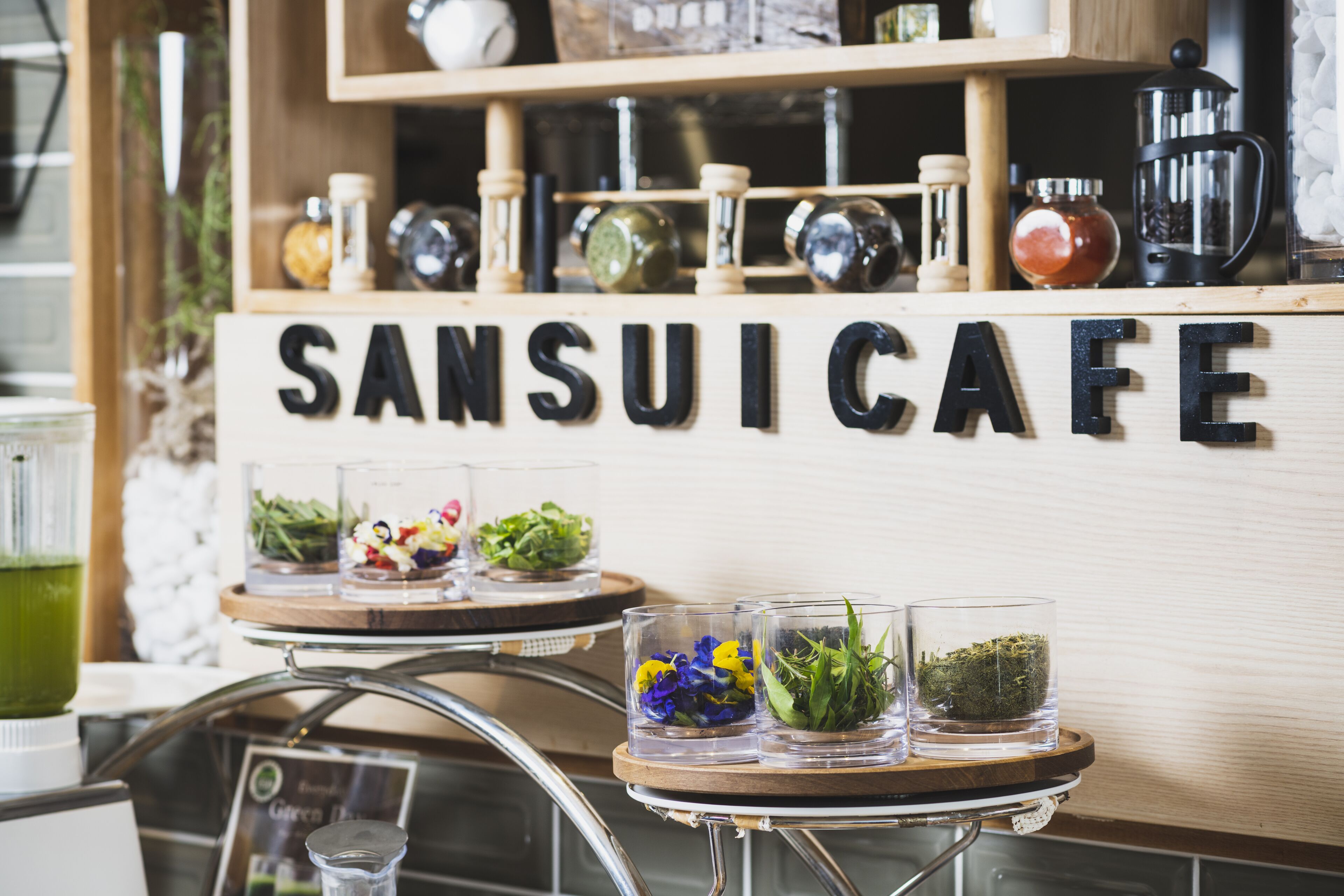 A cafe interior with a sign reading "SANSUI CAFE" and three glass containers with greenery and flowers on a wooden table.