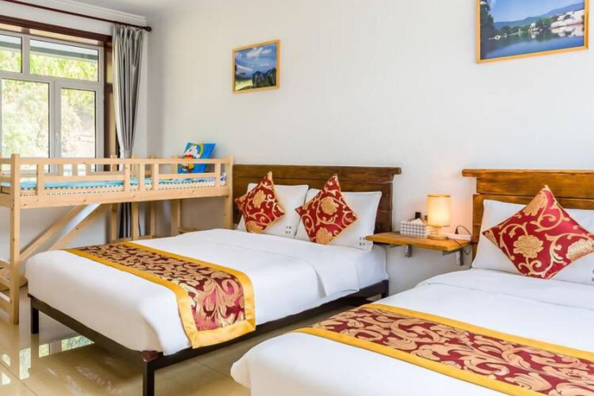A hotel room with two beds, a wooden bunk bed, a window with curtains, a nightstand with a lamp, and a framed picture on the wall.