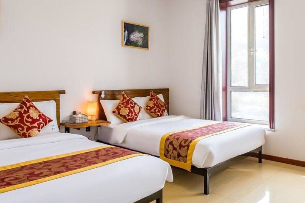 A hotel room with two beds, a nightstand with a lamp, a window with curtains, and a small painting on the wall.