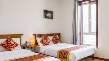 A hotel room with two beds, a nightstand with a lamp, a window with curtains, and a small painting on the wall.