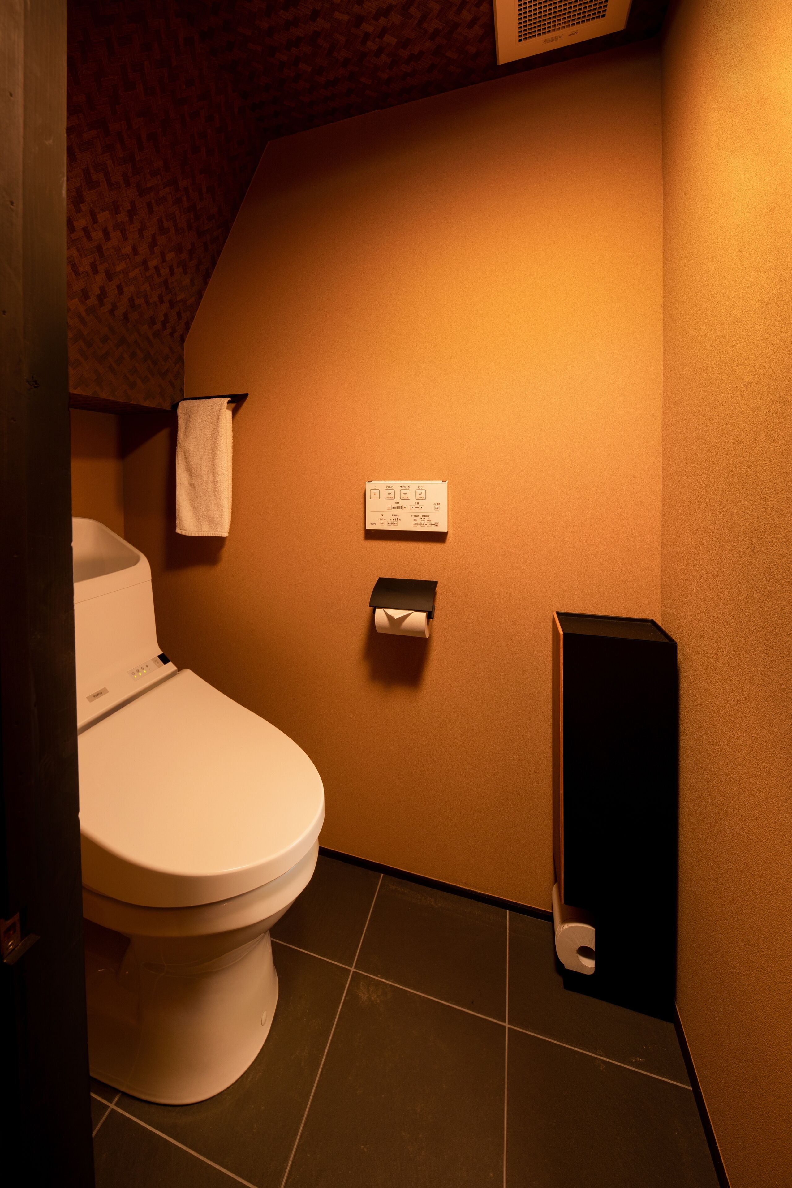 Ein kleines Badezimmer mit Toilette, einem Handtuch an der Wand, einem Papierhandtuchspender und einem schwarzen Lautsprecher.