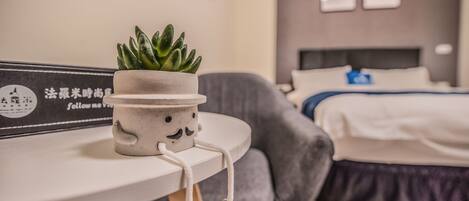 A cozy hotel room with a neatly made bed, a grey armchair, and a small table with a potted plant and a framed sign.