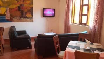 A room with a sofa, a dining table with chairs, a TV mounted on the wall, and a window with curtains.
