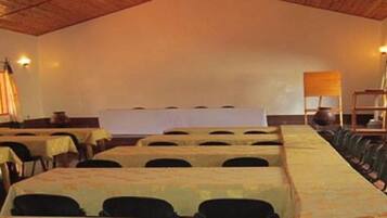 A conference room with rows of tables and chairs, a stage, and windows with curtains.