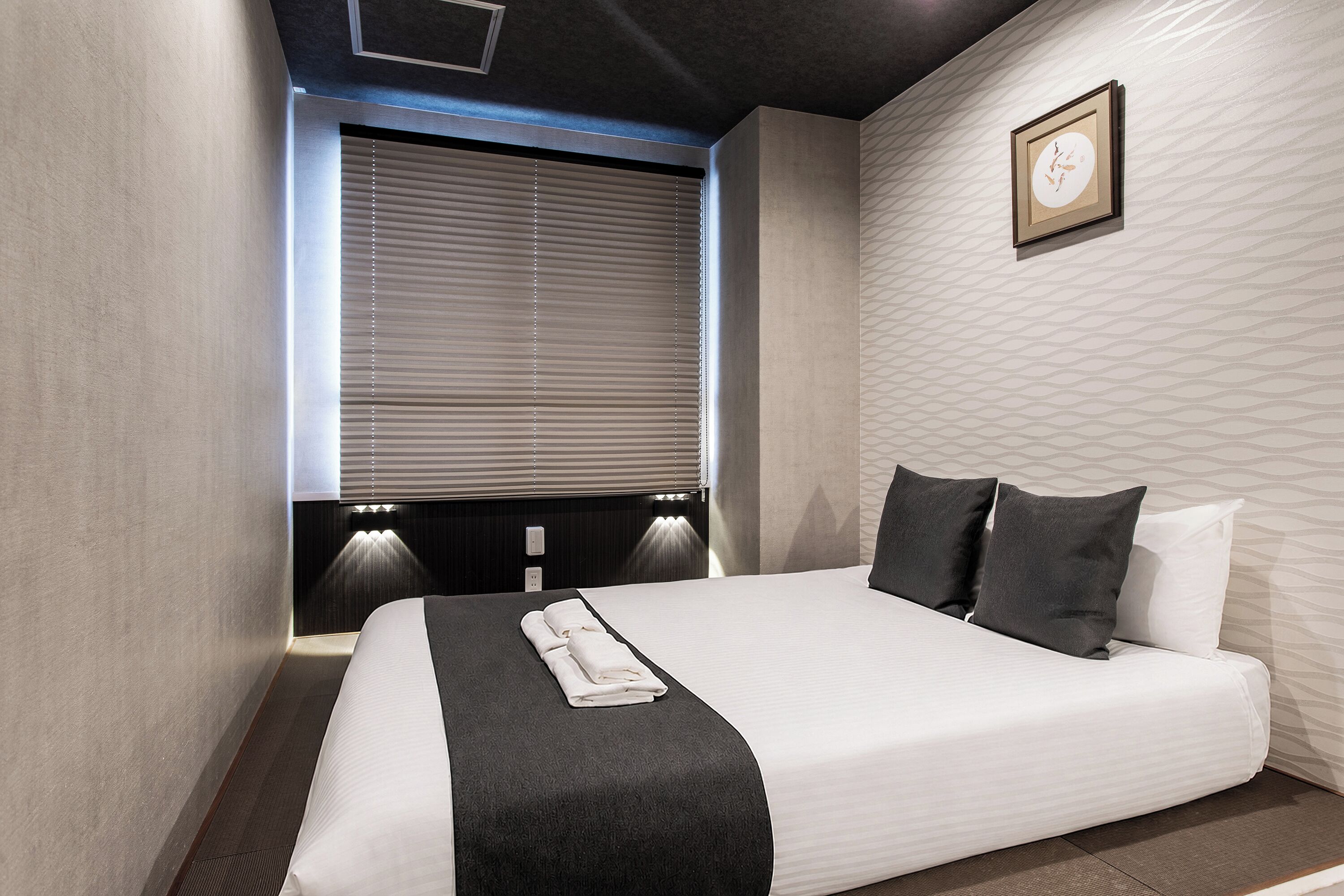 A modern hotel room with a large bed, a window with blinds, a wall-mounted light, and a framed picture.