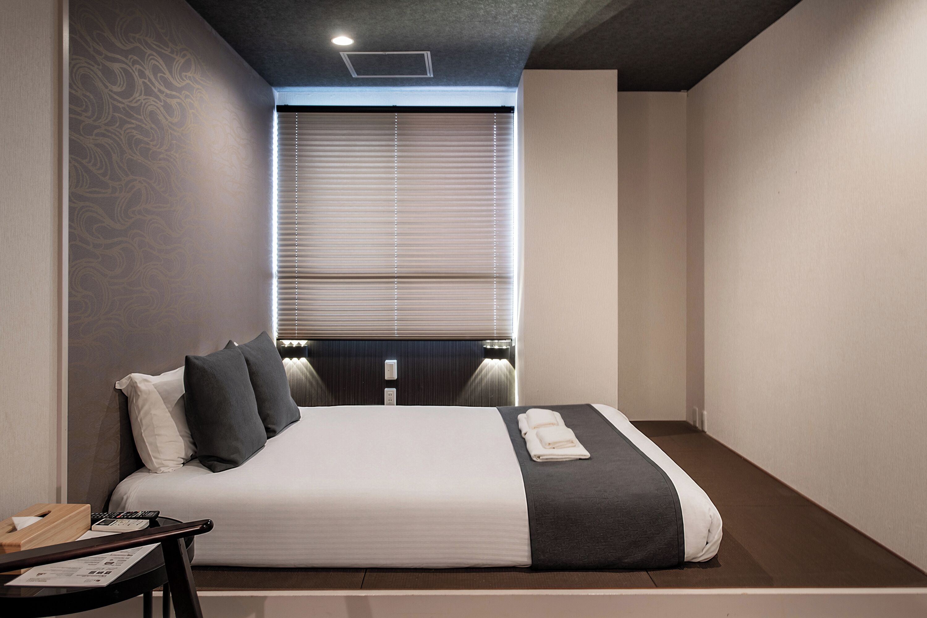 Habitación de hotel moderna con una cama grande, lámparas de noche, un escritorio con teléfono y una ventana con persianas.