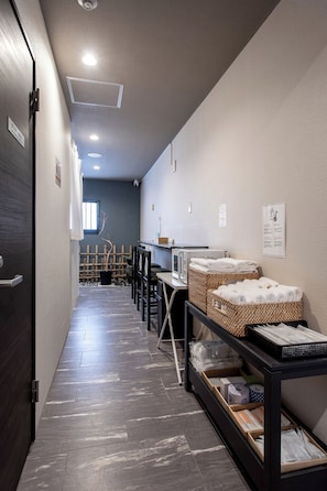 A hotel corridor with a dark wooden door, a white wall with a sign, a microwave, and a table with towels and baskets. - Yuyu Ukiyoe Hotel (Osaka)