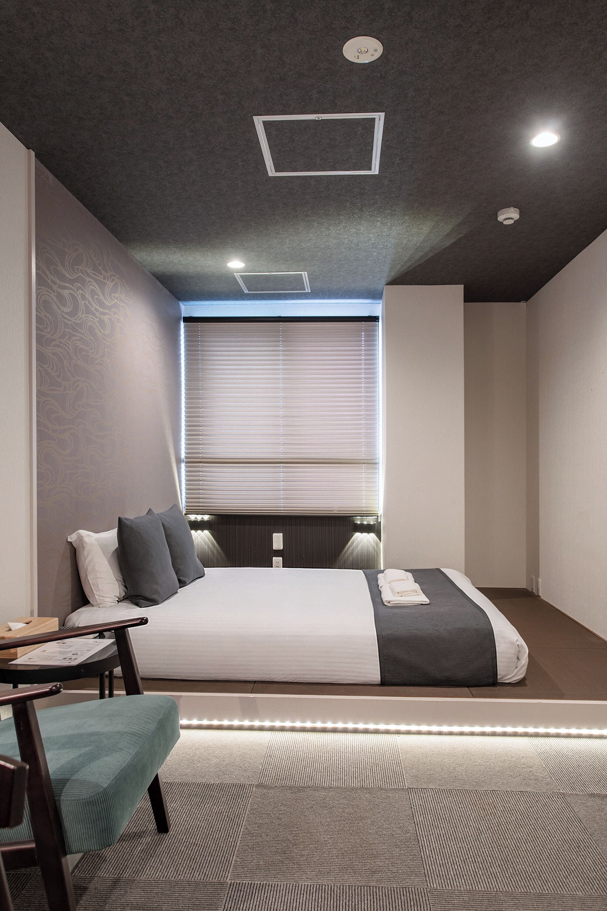 A modern bedroom with a bed, bedside table, chair, and a window with blinds.