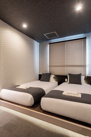 Een hotelkamer met twee bedden, een groot raam met zonwering, een gestructureerde muur en een plafondlamp.