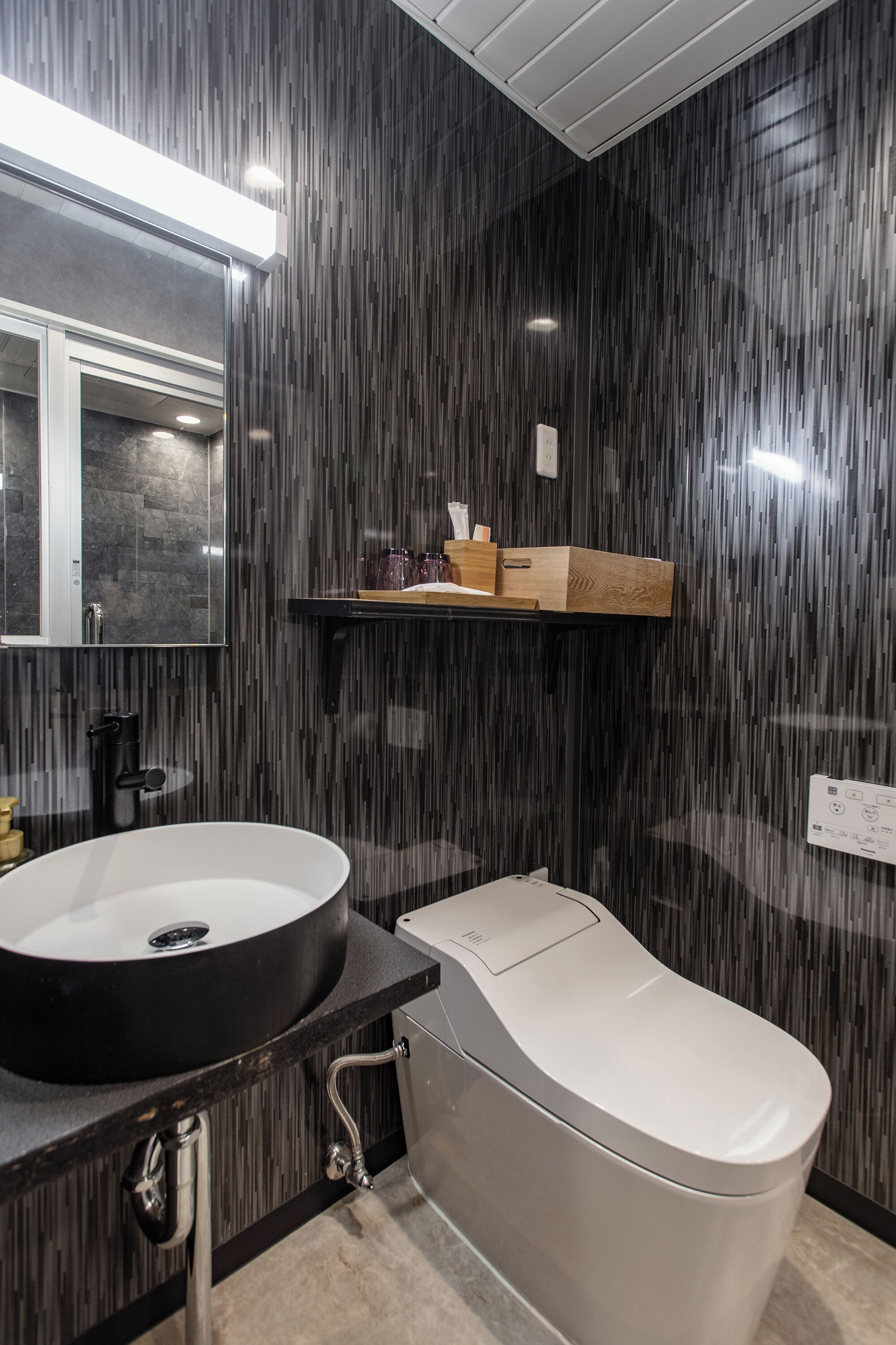 Un baño moderno con un lavamanos negro grande, un inodoro blanco y un estante de madera sobre el lavamanos.