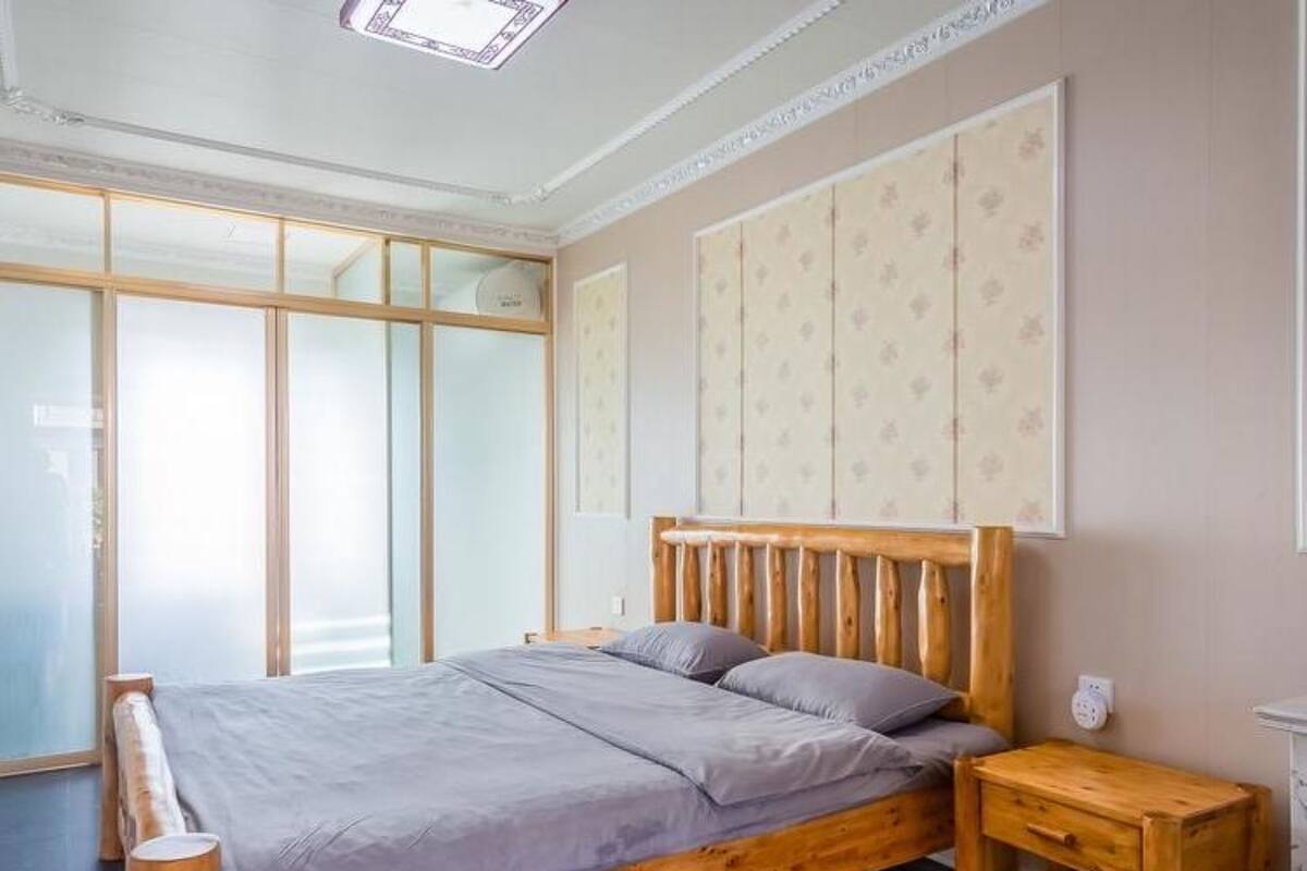 A bedroom with a wooden bed, grey bedding, a small wooden nightstand, and a large window with blinds.