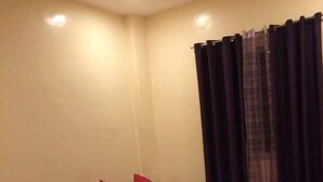 A hotel room with two beds, each with white bedding and red pillows, a small bedside table with a lamp, and a window with dark curtains. - Nathaniel Hotel (Daet)