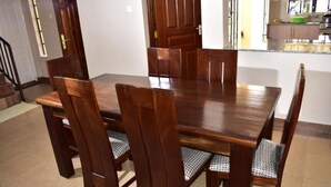 A dining area with a wooden table and chairs, a kitchen in the background, and a staircase leading to another floor. - Valbras Homestay Nairobi (Syokimau)