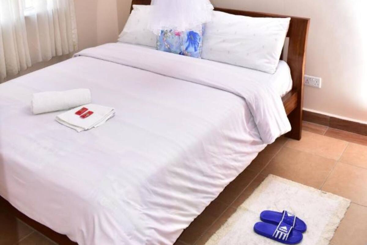 A neatly made bed with a white canopy, a pair of slippers on a rug, and a window with curtains.